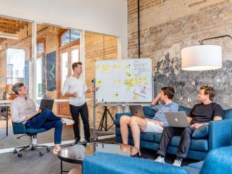 three men sitting while using laptops and watching man beside whiteboard by Austin Distel courtesy of Unsplash.